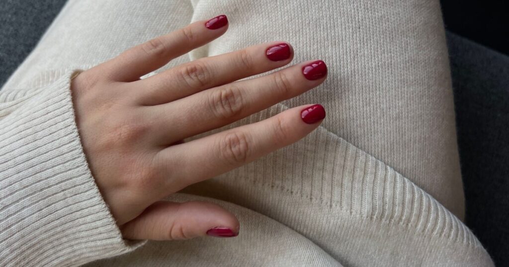 October nails 2026 in deep cherry red on short square nails, resting on a cream knit sweater
